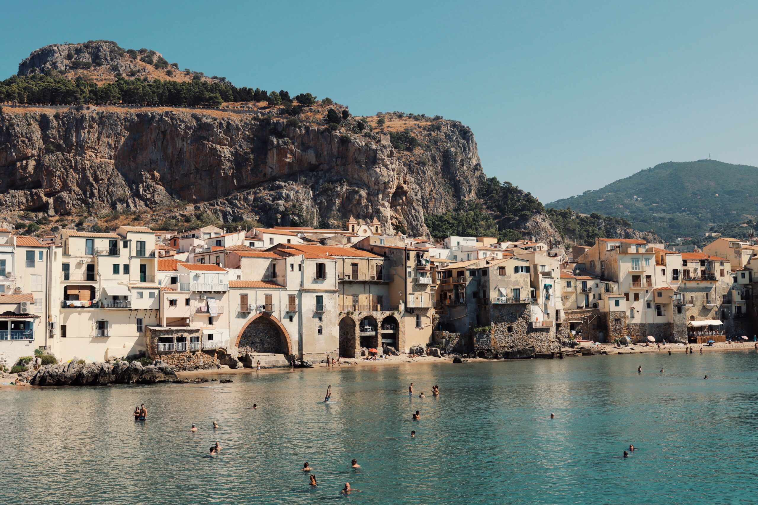 Para perenang menikmati air dengan latar bangunan-bangunan bersejarah di tepi laut Cefalù dan tebing-tebing berbatu di Sisilia, Italia, salah satu lokasi syuting The Odyssey.